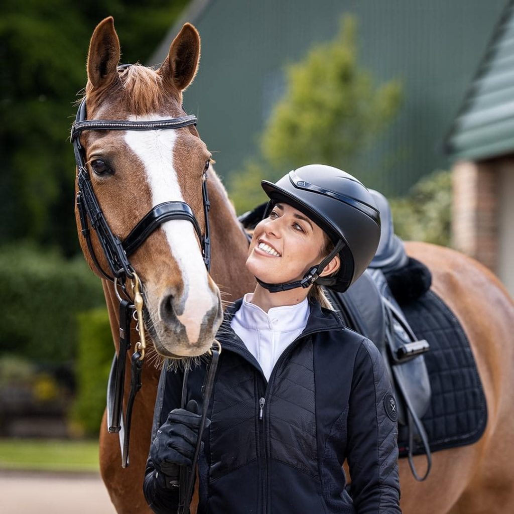 Equestrian in riding gear smiling beside a bridled chestnut horse.